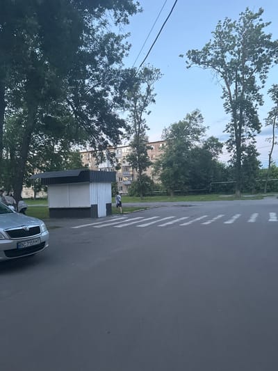 The image depicts a street scene in the city of Гожули, Ukraine. In the foreground, a silver Skoda car is parked on the asphalt, its license plate reading "BC 7159 MT". To the right of the car, a pedestrian crossing is marked with white stripes.
Adjacent to the crossing is a small, kiosk-like structure with a dark roof and white vertical siding, featuring closed white roller shutters on its front. A lone individual, wearing dark shorts and a light-colored shirt with a pattern, is walking away from the kiosk towards the street.
In the background, a multi-story apartment building with numerous windows and balconies stands amidst lush greenery and mature trees. Several other trees are visible throughout the scene, their branches reaching towards a clear, pale blue sky. Power lines stretch across the upper portion of the image. To the left of the kiosk, a group of people, including one person with a stroller, are seated on a bench. The overall environment suggests a typical urban residential area.