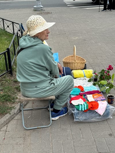 An elderly woman wearing a straw hat and a green tracksuit sits on a folding stool next to a display of handmade knitted items. The items are spread out on a plastic sheet on the ground, including colorful squares, a yellow folded item, and a white lacy cloth. A wicker basket, a potted plant with red flowers, and a cup with a plant are also visible. The scene takes place on a paved area, likely a sidewalk, with a grassy area and a metal fence in the background. The location is identified as Канонерец Снт, Russia.