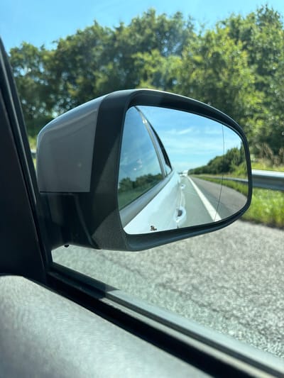 The image shows a side-view mirror of a car on a highway, reflecting the road ahead, trees lining the side of the road, and the sky. The car appears to be in motion, as suggested by the blurred motion of the road and the trees. The scene is bright and sunny, indicating daytime. There are no visible landmarks or signs to specifically place the location within Breda, Netherlands. The mirror itself is a standard car side-view mirror, dark gray in color, with a small sticker or emblem on the lower left corner of the reflective surface. The interior of the car, specifically the door panel and window frame, is visible in the foreground.