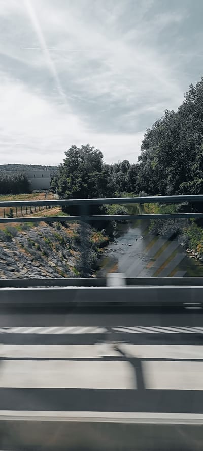 The image captures a scene from a bridge overlooking a river and surrounding landscape, likely taken from a moving vehicle due to motion blur in the foreground. In the foreground, two parallel grey railings run horizontally across the frame, with the lower railing exhibiting motion blur. Below the railings, the blurred surface of a road or pathway is visible, marked with dark and light parallel lines, suggesting speed.
The middle ground features a shallow, meandering river with rocky banks on the left. The river's surface reflects the sky and surrounding trees. On the left bank, a gently sloping area is covered with stones and dry vegetation. Behind the riverbank, a row of metal bollards with a fence separates the area from a field with sparse vegetation and a large, rectangular building with a flat roof, possibly an industrial or commercial structure, situated in the distance. A dense line of trees forms the backdrop, with taller trees to the right and a hill covered in vegetation further in the distance on the left. The sky is overcast with a bright ray of sunlight piercing through from the upper left, creating a slightly ethereal glow. A faint contrail from an aircraft is visible in the sky.
The location identified is Winterbach, Germany. The scene depicts a natural landscape integrated with infrastructure, common in suburban or semi-rural areas of Germany. The time of day appears to be daytime, possibly late morning or early afternoon, given the brightness of the sun. No people are visible in the image, and the focus is on the environment and the man-made structures within it.
