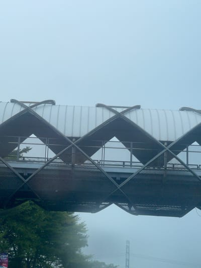 An elevated pedestrian walkway with a corrugated white roof and a dark metal frame is viewed from below against a pale blue, hazy sky. The walkway's structure features a diamond-shaped pattern formed by intersecting metal beams, creating triangular openings. Through these openings, a grey, mesh-like material is visible on the underside of the walkway, and glimpses of trees and the hazy environment beyond can be seen. In the lower left corner, a portion of a sign with the words "Grand Prize" is visible. The overall atmosphere is muted due to the fog or mist. The location is Pakulonan Barat, Indonesia.