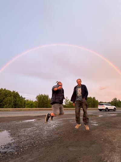 Two men are standing on a gravelly, wet surface next to a road. A white car is parked on the road in the background. Behind the road, there is a line of green trees. A rainbow arcs across the sky. The sky is a light purple color, suggesting it is either sunrise or sunset. The man in the foreground is wearing a plaid shirt and gray pants. The man in the background is wearing a dark jacket over a t-shirt and brown pants. He is looking at the camera and smiling. The man in the foreground is looking down and appears to be holding something in his hands. The overall mood of the image is relaxed and serene.