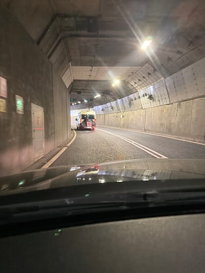 The image captures an interior view of a concrete tunnel, taken from within a vehicle. The perspective looks forward and slightly downward, with a dark, reflective surface on the right side of the frame, possibly a window, which mirrors the scene ahead.
The tunnel's structure consists of concrete walls and ceiling, featuring visible panels and fixtures. Bright artificial lights illuminate the space, with some lights producing noticeable lens flares. Along the left wall, several green lights are visible, possibly signaling or safety indicators.
A roadway or track occupies the center of the tunnel, characterized by a dark, textured surface and distinct double white solid lines that separate two lanes. Ahead, a light-colored vehicle, resembling a maintenance van or small truck, is positioned on the track. Its rear is equipped with prominent red and white diagonal striped reflective markings, and a black cover or equipment is visible.
The foreground on the left shows part of the viewing vehicle's interior or floor, with blurry rectangular lights, one appearing reddish and another greenish. No readable text is discernible anywhere in the image. The scene suggests travel through an enclosed environment, likely a tunnel at night or indoors, within the North Tyneside area of the United Kingdom, possibly a part of a railway or road system.