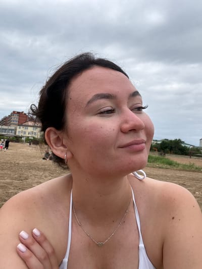 A young woman, with dark hair and fair skin, poses for a selfie on a beach. She is wearing a white bikini and a delicate silver necklace with a heart-shaped pendant. The camera is angled close to her face, capturing her eyes looking upwards and a slight smile. Her skin appears slightly sandy, hinting at the beach environment. In the background, the sand stretches out, leading to a body of water under a cloudy sky. Further in the distance, buildings can be seen, suggesting an urban area adjacent to the beach. The overall mood is relaxed and carefree, typical of a day at the seaside. The time of day appears to be daytime, with the overcast sky indicating a possibly cool or breezy day. The location is identified as Derbent, Russia, which is known for its coastline along the Caspian Sea. There is no visible text in the image.