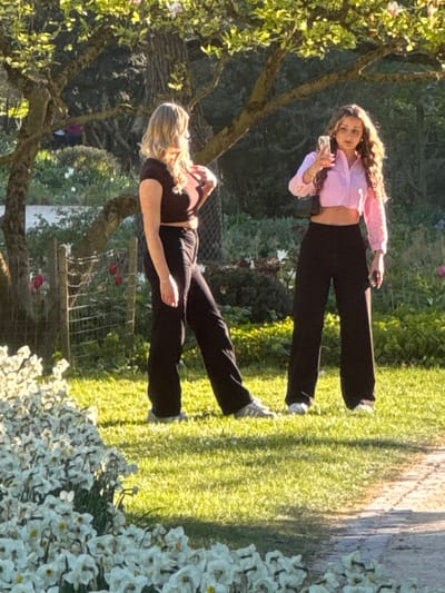 Two young women pose in a garden, situated on a lawn, surrounded by blossoming trees and flowerbeds. The woman in the foreground wears a dark top and black pants. The other woman, located in the background, sports a light pink shirt and dark pants, using a phone. The scene is bathed in sunlight, and it's likely located in Ulm, Germany, though specific city elements are not visible in the frame.