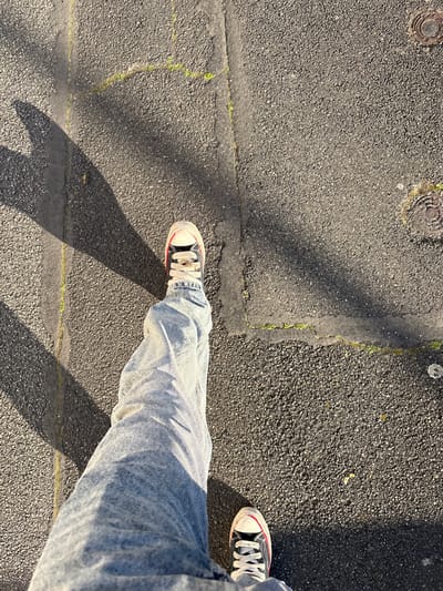 The image depicts a first-person, downward view of a person's lower body while walking on an asphalt surface in Ivry-sur-Seine, France. The individual wears light-wash blue denim jeans and black high-top sneakers with white laces, toe caps, and sole edges, accented with a red stripe. The left leg is extended forward, while the right foot is grounded. The asphalt pavement is dark gray, granular, and exhibits several linear cracks and seams, some of which contain green moss or vegetation. Two circular, textured metal utility covers are visible on the right side of the frame. Strong, distinct shadows are cast from the left across the pavement, indicating direct sunlight.
