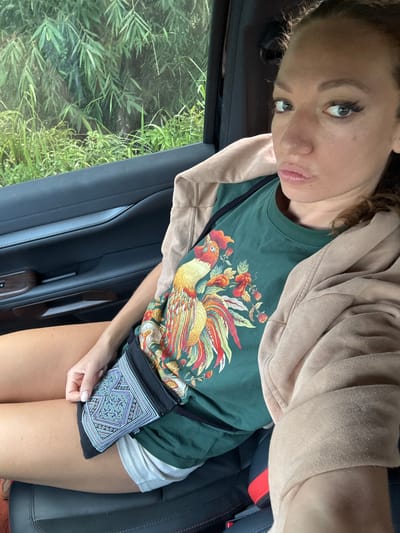 A fair-skinned female is posing for a selfie inside a vehicle. She is wearing a tan hooded sweatshirt over a dark green t-shirt featuring a large rooster design. She is also wearing white shorts. A small, black patterned bag hangs across her body. The woman has brown hair and is looking toward the camera with a slight pout. Outside the vehicle's window, green foliage is visible. The vehicle appears to be a car, judging by the interior door panel and seat. The specific location of Hoàng Su Phì Commune, Vietnam cannot be verified from the image.
