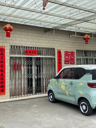 This image depicts a festive scene outside a home in Dujiangyan City, China, decorated for a celebration, likely a wedding. A light green electric car, adorned with Pokémon stickers featuring Pikachu and lightning bolts, is parked in front of a building. The building has a tiled facade and is covered by a transparent corrugated roof supported by metal beams. Red lanterns hang from the roof, and strings of small red lights are strung across it.

Decorative red banners with Chinese characters are affixed to the wall. One banner on the left reads "龍鳳呈祥幸福美滿好姻緣" (Dragon and phoenix bring prosperity, happiness, and a good marriage). Above the metal security gate of the entrance, a horizontal red banner reads "合好年百" (Harmonious and good for a hundred years). To the right of this, a large red Chinese character "囍" (double happiness), commonly used for weddings, is displayed. The security gate is closed, and behind it, curtains with a floral pattern are visible. The ground in front of the building is paved with concrete. The overall impression is one of a joyous occasion being celebrated.