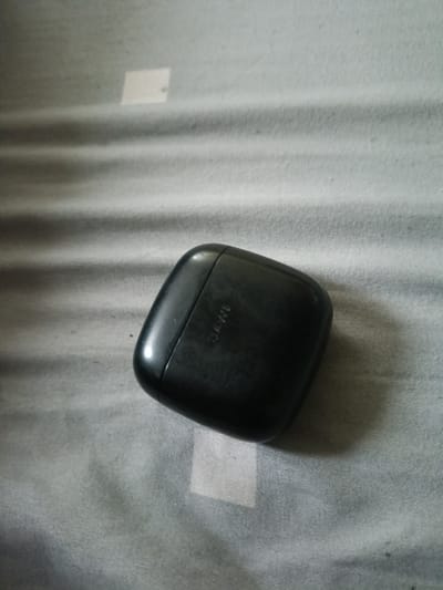 A black wireless earbud charging case is placed on a gray, textured surface. The case is oriented with its lid facing slightly upwards and to the left. The word "SAWE" is embossed on the lid. The background appears to be a fabric with horizontal lines or folds, possibly a bedspread or a curtain. There are no people or other objects visible in the image. The image was taken indoors.