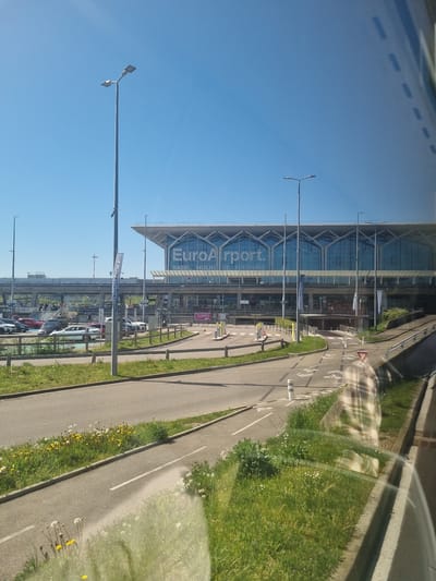 The image shows EuroAirport Basel-Mulhouse-Freiburg in Saint-Louis, France. The airport building features a glass facade with the name "EuroAirport" displayed prominently. A road runs alongside the airport, with bicycle lanes marked. Streetlights line the road. Cars are parked in a lot adjacent to the road. The sky is clear and blue. Vegetation, including grass and flowers, grows along the roadside. The image appears to be taken from within a vehicle, as part of the window is visible.