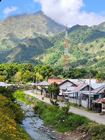 The image shows a view in Sembalun Bumbung, Indonesia, featuring a foreground stream choked with trash alongside a road running through a small village. Houses with corrugated metal roofs line the street. A person is riding a motorcycle on the road. In the background, a large, forested mountain with a tall communications tower rises, its peak partially obscured by clouds. The sky is blue with scattered clouds. Yellow wildflowers grow along the banks of the stream.