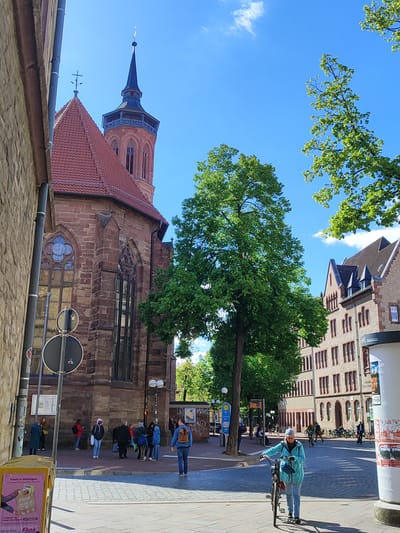 The image depicts a street scene in Göttingen, Germany, showing the exterior of St. Jacobi Church and surrounding buildings. A group of people is gathered near the church entrance, while another individual walks toward the camera, pushing a bicycle. Cobblestone pavement covers the street. The sky is bright blue with some clouds. Street signs and advertisements are also visible. The scene is bathed in daylight.
