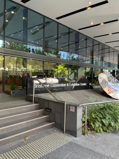 The image captures the exterior entrance of the Brisbane Convention & Exhibition Centre in South Brisbane, Australia, during what appears to be a bright day. The main subject is a modern, multi-story building featuring extensive glass panels that reflect the sky, trees, and other architectural elements.

A prominent black pillar to the right of the entrance features the white, vertically oriented text: "Brisbane Convention & Exhibition Centre". To the right of this pillar, a set of outdoor stairs with a metal handrail leads down from the entrance level. The stairs and surrounding ground are made of grey tiles, with tactile paving visible on the sidewalk area, suggesting accessibility features.

To the left of the stairs, there's a small garden bed with lush green plants. Above this greenery and near the building's facade, a distinctive, colorful, quilt-like art installation or umbrella structure is partially visible. Further to the right, a "NO SMOKING" symbol is affixed to the grey wall beside the stairs. No people are visible in the scene, and no specific activity is taking place, but the area is clearly designed as an entryway for a large public venue. The overall impression is one of a clean, well-maintained, and modern urban environment.