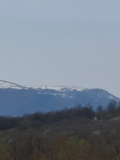 A landscape image depicts a mountain range on the right side with snow on the peak. The left side of the image is a light blue sky. The terrain at the base of the mountains is forested. The image appears to be taken during daylight. There is no visible evidence to indicate that this image is in Komarevo, Bulgaria.