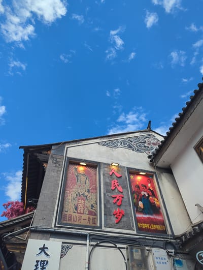 This is a low-angle, eye-level shot of a building facade in Dali City, China, under a bright blue sky with scattered white clouds. The building features traditional Chinese architectural elements, with a grey tiled roof and ornate decorative patterns on the walls.

The facade prominently displays two large, framed posters. The poster on the left depicts a historical figure, possibly Mao Zedong, raising his arm in victory, surrounded by a crowd of people. The text beneath translates to "Long live the motherland and its rivers and mountains." The poster on the right shows three young people in revolutionary attire, holding a sickle and a rifle. The text below reads, "Long live the People's Republic of China."

Below these posters, a white sign with black Chinese characters is visible, which appears to read "大理" (Dali). Electrical conduits and a junction box are mounted on the wall beneath the signs. To the left of the main facade, a branch with bright pink blossoms adds a touch of color to the scene. The right side of the image shows the edge of an adjacent building with white walls and a dark tiled roof.