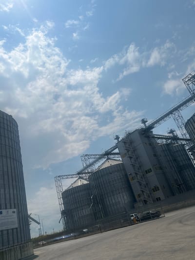 This is an eye-level, wide shot of industrial grain silos under a bright, partly cloudy sky. The orientation of the image is rotated 90 degrees counter-clockwise. The scene depicts a large-scale agricultural or industrial facility.
The primary subjects are the towering grain silos, constructed from metal, with intricate external scaffolding and access platforms. These structures dominate the frame, reaching towards the sky. Visible on the silos are numerous windows and what appear to be ventilation systems. In the foreground, a long, grey, corrugated metal building also suggests an industrial or storage purpose. A paved area, possibly a road or loading dock, runs alongside the buildings on the right.
A car is visible parked near the silos, and some streetlights are present, indicating infrastructure. The weather appears to be clear with scattered white clouds, suggesting it is daytime. The sun is casting some light and glare on the scene. There are no people clearly visible.
A sign is affixed to the foreground building with text and logos, including what appears to be the Bulgarian flag and European Union symbols. The text on the sign is partially obscured and too small to read clearly, but it seems to be informative, possibly related to the project or its funding. The location is identified as Razdelna, Bulgaria. The overall impression is one of an operational industrial site.