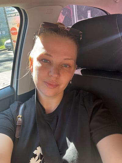 A young woman with blonde hair and freckles is taking a selfie in the passenger seat of a car. She is wearing a black t-shirt with a floral print and has sunglasses on top of her head. The car's interior is visible, including a black headrest and a seatbelt. Outside the car window, a street with buildings and trees can be seen. A billboard with Russian text is also visible. The image appears to be taken on a sunny day.