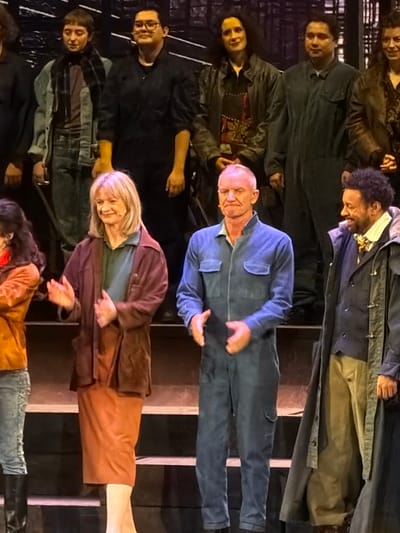 A group of ten individuals is captured on a stage, likely within a theater in Boulogne-Billancourt, France. The scene suggests a curtain call or group acknowledgment, with performers illuminated by stage lighting against a dark, abstract backdrop that hints at urban or industrial structures.
In the foreground, from left to right, four individuals are visible from the waist up. A partially visible woman on the far left wears an orange-brown jacket. Next, a blonde woman in a teal shirt, rust-brown jacket, and matching skirt is actively clapping with a smile. Central to the foreground is a fair-skinned man with short grey hair, identified as Sting, wearing a long-sleeved blue utility jumpsuit. He stands with a neutral expression, hands slightly raised and open. To his right, a dark-skinned man with a beard and dark curly hair smiles broadly, looking towards Sting. This man is dressed in a light shirt, yellow bow tie, dark vest, khaki trousers, and a long dark coat.
Behind this front row, on a slightly elevated dark platform, six more individuals are visible. They are variously dressed in casual or costume attire, including a woman with short dark hair and a plaid scarf, a man with glasses, and others in dark jackets or jumpsuits. Their expressions range from neutral to smiling, and they generally face forward, acknowledging an unseen audience. The stage itself features visible dark steps or tiered platforms.