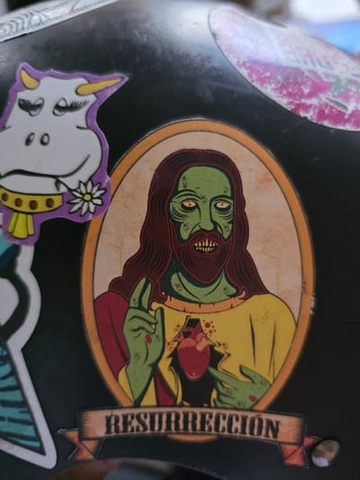 The image displays a close-up of multiple stickers applied to a curved, black surface, likely a helmet or similar object, exhibiting minor scratches and dust. The central and dominant sticker is an oval-framed illustration of a figure resembling Jesus Christ, but depicted with zombie characteristics. This figure has green skin, dark red hair and beard, pale yellow eyes with dark pupils, and an open mouth revealing sharp, exposed teeth. Bloodstains are visible on the figure's face and hands. The figure wears a yellow tunic and a red cloak. Its left hand is raised with two fingers extended, showing a red mark (stigmata) with dripping blood. The right hand points to an anatomical heart, rendered in red, visibly exposed through a jagged opening in the figure's chest, surrounded by red splatters. Below this oval sticker, a banner with faded brown shading reads "RESURRECCIÓN" (Spanish for "RESURRECTION") in a bold, distressed font. To the top-left of the zombie Jesus sticker, a cartoon sticker of a white cow is visible. This cow has purple horns and outline, closed eyes with long eyelashes, a small smile, and holds a white flower with a yellow center in its mouth area, while wearing a yellow and brown collar. A partially visible, faded sticker with pink and white patterns is located in the top-right corner of the black surface. The specific location in the city of Villa San Andrés, Argentina, cannot be determined from the visual information provided in the image.