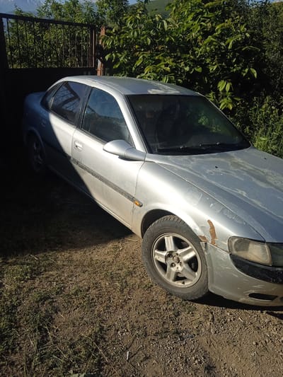 An exterior, eye-level, medium shot captures a silver sedan parked on a dirt and grass surface under a bright, sunny sky. The car is positioned at an angle, facing right, with its front right side prominently featured. The vehicle exhibits signs of wear and tear, including peeling paint and rust on the fender. Overgrown green foliage and a dark metal gate are visible in the background. The scene appears to be located in a rural or semi-rural area, possibly in or near Aznvadzor, Armenia, given the presence of the car, gate, and natural surroundings. No people are visible in the image.