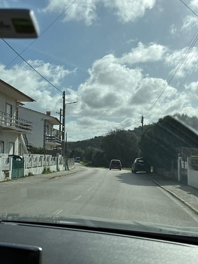 The image depicts a rural or semi-urban road scene in Mafra, Portugal, observed from the interior of a vehicle looking forward through the windshield. The foreground shows a dark dashboard. The asphalt road features faded double white lines marking the center, and curves gently to the left in the distance. On the left side of the road, two multi-story white residential buildings with terracotta roofs and balconies are visible behind a white picket fence with a green metal gate. A utility pole with multiple streetlights stands near the buildings, with overhead power lines traversing the sky. On the right side of the road, a dark-colored SUV is parked off the asphalt, partially obscured by dense green vegetation. Further down the road, a red hatchback vehicle is visible, appearing to move away from the viewer. In the background, beyond the immediate roadside, a hilly landscape covered in green trees and bushes extends, with a white bus discernible on the road in the distance. The sky above is light blue with scattered white cumulus clouds. A blurred rectangular object, possibly a sun visor or sticker, displaying red and green elements, is visible at the top edge of the frame, originating from inside the observer's vehicle.