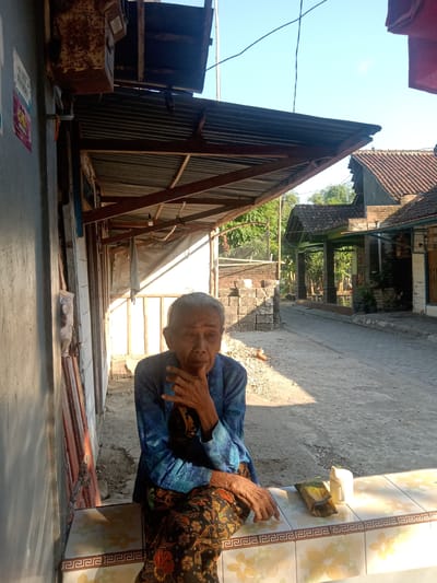 An elderly person with grey hair and weathered skin, wearing a blue patterned shirt and a batik skirt, sits on a tiled ledge outdoors. Their left hand is raised to their chin, and their right hand rests on their lap. In front of them on the ledge are two items: a wrapped food item, possibly a banana leaf-wrapped rice cake, and a small white container. The background shows a street with buildings and a clear sky. The setting appears to be a residential area in Jumput, Indonesia. The architecture suggests a tropical climate with open structures and tiled roofs. A corrugated metal overhang provides shade over the area where the person is seated. The ground is a dirt and gravel pathway.