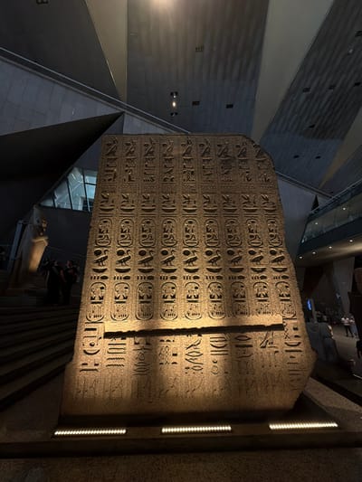 The image displays a large, weathered stone obelisk covered in intricate hieroglyphic carvings. The obelisk stands upright within a dimly lit, modern museum or exhibition space, with angular architectural elements and spotlights illuminating the ancient artifact. The lighting casts shadows across the hieroglyphs, highlighting their texture and depth.

In the background, glimpses of a contemporary structure are visible, including angled walls, ceiling elements, and what appear to be staircases or ramps. A faint reflection on a glass surface suggests an indoor environment. Near the base of the obelisk, a light-colored statue of a seated figure, possibly a pharaoh or deity, is partially visible. A few people are also discernible in the background, their presence indicating that this is a public space, likely a museum exhibit.

The overall atmosphere is one of historical reverence and modern presentation. The contrast between the ancient obelisk and the contemporary architecture creates a sense of timelessness, juxtaposing past and present. The hieroglyphs, the primary subject, are dense and organized in rows, demonstrating the skill of ancient artisans. The scene appears to be set indoors, as evidenced by the lighting and architectural style, and likely at night or in low-light conditions to emphasize the exhibit. No specific weather or time of day can be determined beyond it being an indoor, lit environment. The location context provided is Kafrat Nassar, Egypt, which would suggest this is an Egyptian artifact displayed within that region. No visible text other than the hieroglyphs on the obelisk is clearly legible.