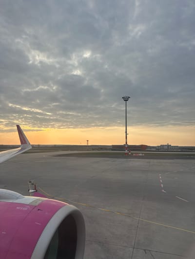 The image depicts an airport tarmac scene at either sunrise or sunset, viewed from the window of a stationary aircraft at Ferenc Liszt International Airport (BUD) in Vecsés, Hungary. The sky is largely overcast with textured grey clouds, transitioning to a bright orange-yellow glow on the horizon. In the lower left quadrant, a portion of a white aircraft wing with a red/pink winglet, bearing the text "wizzair.com," is visible. Below the wing, the forward section of a pink and white aircraft engine nacelle with a grey intake is prominent, displaying "NO STEP" stenciled text. A small white ground support equipment vehicle is positioned on the tarmac near the engine, connected to the aircraft by a yellow line. The tarmac itself is a vast grey concrete surface with various white dashed lines and solid yellow markings. In the mid-ground, a tall, slender floodlight tower with a cluster of lamps at its apex and red and white striped markings on its lower section stands centrally. In the distant background, silhouetted against the bright horizon, are a slender air traffic control tower with a rounded top and several airport terminal or hangar buildings featuring orange and grey facades. No individuals are visible in the scene.