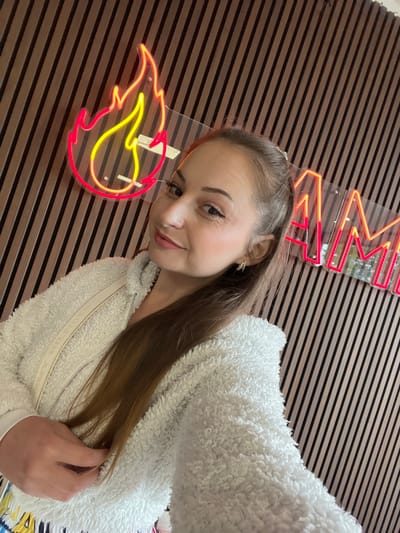 A waist-up, selfie-style photograph features an adult female posing indoors against a diagonally striped wall. The woman, with light skin and long, straight brown hair partially pulled back, looks directly at the camera with a slight smile. She is wearing a textured, off-white, sherpa-style full-zip jacket. Her left ear is visible, adorned with a small, possibly crystal earring. Her right hand is partially visible at the bottom left, resting on her jacket. In the background, mounted on clear acrylic panels against the wall, are two neon signs: a prominent red and yellow flame icon on the left, and partially visible red neon lettering to the right that appears to spell "AM...". The wall itself is composed of alternating light and dark brown diagonal stripes, resembling wood paneling. The scene is located in Oostham, Belgium.