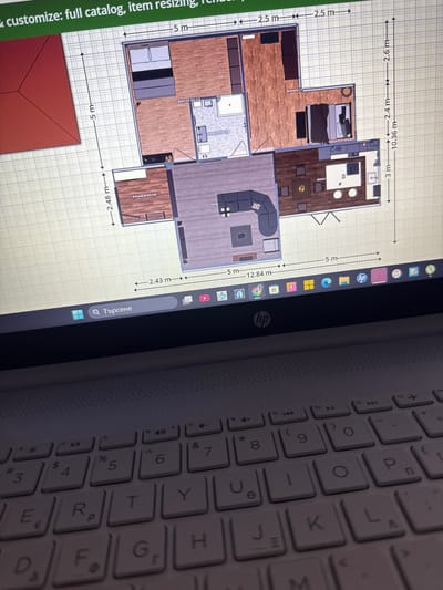 The image shows a close-up view of a laptop screen displaying a floor plan, likely for interior design or real estate purposes. The floor plan is detailed, showing rooms with dimensions labeled in meters, such as "Bedroom (18.465 m2)", "Kitchen", and "Entryway (6.023 m2)". Furniture is depicted within the rooms, including a bed, sofas, a dining table with chairs, and a kitchen island with stools. The floor plan is overlaid on a grid.
To the right of the floor plan, a portion of the laptop's keyboard is visible. The keyboard is white and features Cyrillic characters, indicating the likely language setting. The screen also shows icons for various applications along the side, including "hp" logos, suggesting it's an HP laptop. A search bar with the text "Q Търсене" (which translates to "Q Search" in Bulgarian) is also visible.
The overall scene is an indoor setting, likely a home office or living space, where someone is actively using the laptop for design or planning. The lighting appears to be artificial, casting a warm glow on the scene, and no specific time of day or weather conditions are discernible. The image is focused on the laptop screen and keyboard, with the rest of the environment blurred out.