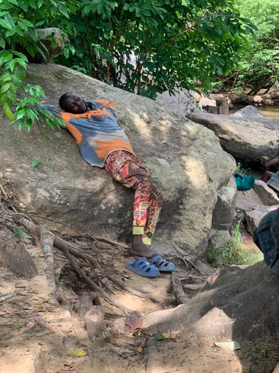 A man is lying on his back on a large rock, propped up on his elbows, in Egbeda, Nigeria (latitude: 7.119, longitude: 5.362). He is wearing a long-sleeved, orange and gray shirt with a faded pattern and colorful patterned pants. His bare feet are wearing blue sandals. There is a large, gray rock behind him, along with a small blue bucket to the right of the frame. The man's eyes are closed and he appears to be sleeping or resting. There is dense foliage and vegetation in the background, as well as a small stream. The scene is partially shaded from the sun by trees. The ground is sandy and there are large roots protruding from the soil, likely from the nearby trees.