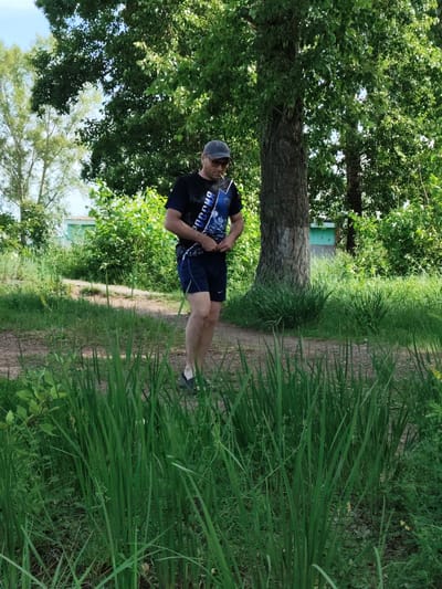 A man stands in a grassy area, wearing a dark t-shirt with an image and text that appears to be in Cyrillic, dark blue shorts, a baseball cap, and sunglasses. He is holding a flat, rectangular object, possibly a book or a clipboard, in his left hand, and adjusting the front of his t-shirt with his right hand. The setting is outdoors, with a dirt path leading past him. Tall green grass and foliage are prominent in the foreground, and a large tree with a thick trunk stands behind the man. The background shows more trees and some greenery, with a hint of a light-colored building in the distance. The lighting suggests it is daytime, likely sunny, based on the shadows and the brightness of the scene. The man's expression is not clearly visible due to his sunglasses, but his posture suggests a casual stance. The text on his t-shirt reads "РОССИЯ", which translates to "RUSSIA". The overall scene depicts a relaxed outdoor moment in what appears to be a rural or park-like environment in Raikov, Russia.