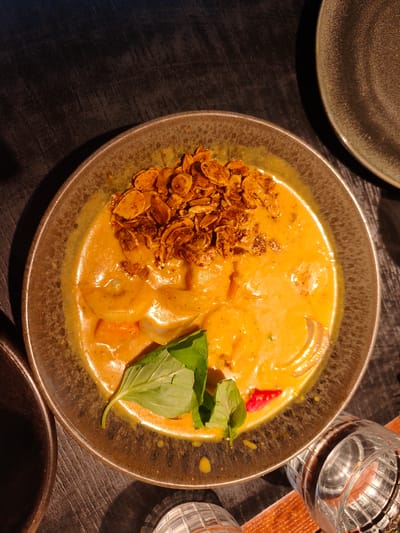 The image displays a close-up, top-down view of a bowl of curry on a dark, textured wooden table. The bowl is a speckled brown and grey ceramic, filled with a vibrant orange curry. Toppings include a generous scattering of what appear to be fried shallots or garlic chips. Visible within the curry are pieces of food, including what seem to be dumplings or pasta, and a few bright red slivers, possibly bell pepper. A fresh green basil leaf is placed on the side of the bowl. To the right of the bowl, two clear, patterned glasses are partially visible, filled with a clear liquid, suggesting drinks are present. In the background, another dark plate is positioned to the right, and a section of a dark surface occupies the upper left. The lighting suggests an indoor setting, possibly a restaurant, with shadows cast across the table. The image does not contain any people or provide information about the specific location within Bnei Dror, Israel.
