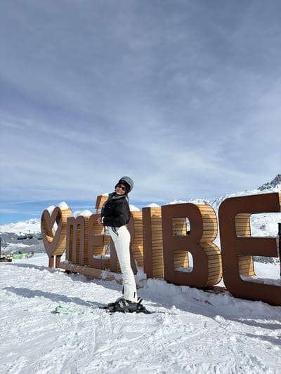 The image displays an outdoor winter scene at the Méribel ski resort, located within the Three Valleys ski domain, France, which is accessible from Saint-Martin-de-Belleville. A single female individual stands centrally, posing for the camera on groomed snow. She is equipped with a dark grey ski helmet, dark goggles positioned on the helmet, a black puffer jacket, white ski pants, and dark ski boots. One ski boot is attached to a black ski binding on a ski, while a second ski with a green base and black top lies flat on the snow to her left. Her head is turned towards the viewer, displaying a smile. To her left, a large, free-standing sign spells "❤️ MERIBEL" in dark, horizontally slatted letters, partially topped with snow. In the background, a ski lift with yellow chairs ascends a snow-covered slope, with a partially visible green sign below it bearing the text "DEPART". Distant snow-covered mountain ranges are visible under a bright, partially cloudy blue sky.