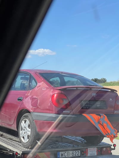 The image shows the rear of a red Toyota Avensis sedan being transported on a car carrier trailer. The car's paint is visibly faded and peeling, particularly on the rear quarter panels. A bright orange and grey high-visibility vest is draped over the rear bumper. The car has a license plate that appears to read "WCB-622 H". A business name and phone numbers are visible on a sticker on the rear of the trailer. The background suggests an outdoor, possibly rural setting, with a blue sky and scattered clouds. The lower left portion of the image is obscured by a dark, angled shadow, likely from another vehicle or structure. The scene appears to be in Podivín, Czechia, based on the provided location context. There are no visible people in the image.