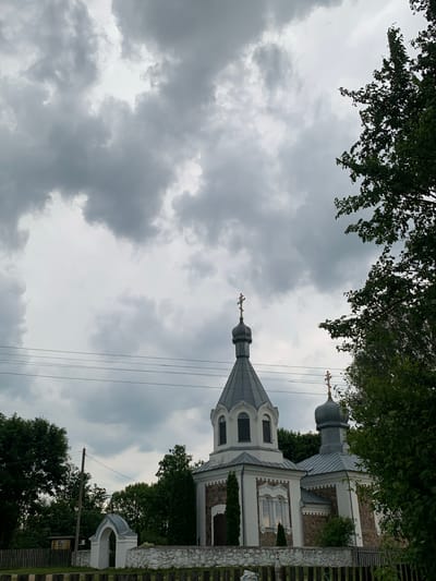 The image depicts a church with multiple domes against a cloudy sky, with trees and power lines in the foreground. The church is white with stone accents and features several spires topped with crosses. To the left of the church, there is a white arched gateway and a wooden fence. The overall scene suggests a rural or semi-rural setting in Орлеўскі сельскі Савет, Belarus.