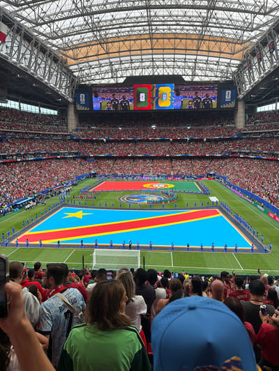 This media file captures a lively scene at a soccer stadium, likely in Houston, United States of America, during an event. The image is oriented vertically, showing a wide view of the stadium and the field.
The main subjects are the numerous people filling the stands, dressed in red and white attire, creating a vibrant backdrop. On the field, a large replica of the Democratic Republic of Congo's flag is prominently displayed. Lined up along the flag are individuals in matching blue uniforms, presumably players or participants in the event.
The scene appears to be a pre-game ceremony or introduction, with the flag display and uniformed individuals suggesting a formal occasion. The stadium's distinctive architecture, with its intricate metal framework, is visible in the background. Large screens are mounted on the stadium's structure, showing images of people in uniform. One screen displays the FIFA World Cup 2026 logo along with other graphics.
While the time of day is not explicitly stated, the bright lighting suggests it is daytime. The atmosphere seems excited and celebratory, judging by the crowd's density and engagement.
Visible text includes "FIFA WORLD CUP 2026" on a banner, as well as various sponsor logos such as "Coca-Cola" and "HYUNDAI". Some individuals in the crowd are holding up their phones, possibly to capture the moment. The overall impression is one of a significant sporting event, filled with anticipation and national pride.