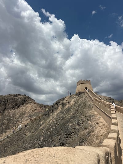 The image captures a section of the Great Wall of China, specifically the Jiayu Pass in Jiayuguan, China. The perspective is from a low angle, looking up towards a watchtower situated on a rocky, arid hilltop. The watchtower, constructed of tan bricks, features crenellated battlements. A long, winding pathway with its own battlements leads up to the tower, with several tourists in the distance. Some tourists are also visible on a separate staircase winding up a steeper, more rugged section of the hillside to the left. The landscape is characterized by dry, rocky terrain with sparse vegetation. The sky above is a mix of deep blue and dramatic, heavy grey clouds, suggesting an impending weather change.