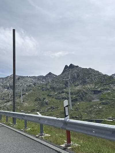 This image depicts a mountain scene, likely in Airolo, Switzerland, given the context. The foreground is dominated by a road barrier made of metal poles and a silver rail. To the left of the barrier, a long, dark metal pole extends upwards from the bottom left corner of the frame and disappears near the top. The background features a steep, rocky mountain covered in patches of green grass and small patches of snow. The sky is overcast with soft white clouds, suggesting a cool or temperate day.
Several pieces of infrastructure are visible along the roadside and on the mountain. A white and grey object with a dark rectangular panel is mounted on a pole near the road barrier. Further up the mountain, the faint outlines of what appear to be power lines or a ski lift system can be discerned. The overall impression is one of a remote, natural landscape with some human intervention for infrastructure. There is no visible text in the image. The activity suggested is simply the presence of a road, likely a scenic route, through the mountains. The weather appears to be overcast, and the time of day is indeterminate due to the lack of distinct shadows.