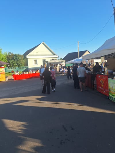 The image depicts an outdoor market scene on a sunny day, likely in Цімкавіцкі сельскі Савет, Belarus. In the foreground, the asphalt road is partially covered by the shadows of trees and structures. Several people are present, browsing and interacting at market stalls.
On the left side of the image, a yellow food stand is visible, with a red table displaying various items, possibly crafts or goods, behind it. A group of people are gathered around this area, with some appearing to be vendors and others customers. Further back, a light-colored residential house with a peaked roof is situated.
Towards the right side of the image, several white tents or canopies are set up, indicating more market stalls. One stall appears to be selling food, with a person behind a counter and bottles of beverages visible. Another stall features a red banner with Cyrillic lettering and imagery. People are seen standing and walking between these stalls. The background reveals more buildings and trees, characteristic of a rural or semi-rural setting. The sky is clear and blue, suggesting fair weather conditions.