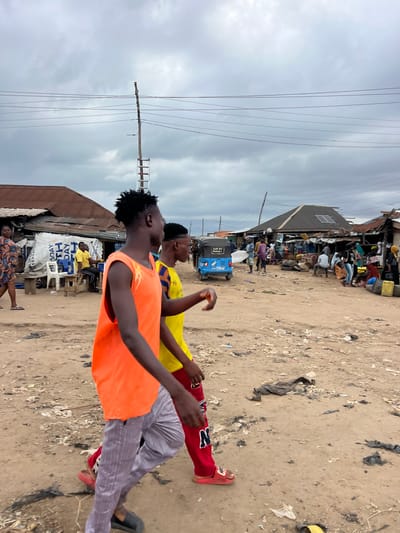 The image captures a street scene in Oko Amakom, Nigeria, under an overcast sky. In the foreground, two young men are walking from left to right. The man on the left wears a bright orange sleeveless t-shirt and light-colored striped pants. The man to his right wears a yellow t-shirt with blue and red stripes on the sleeves, and red pants with a white "N" logo. They are walking on a dirt path littered with debris, including plastic bags and torn fabric.
The background reveals a bustling market environment. Structures with corrugated metal roofs line the street, some appearing makeshift and worn. Several individuals are engaged in various activities: standing, sitting, and walking. A blue auto-rickshaw is parked on the left side of the street, displaying text that includes "NAM GAI DAMARI". Power lines are strung across the sky, supported by wooden poles. The overall impression is a busy, somewhat unkempt urban setting.