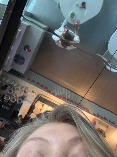 A woman's face dominates the right side of the frame, her gray hair partially obscuring the scene. The image reflects in a surface, showing a table with dishes and a person in a white shirt holding a phone. Also reflected is a cabinet with circular elements, along with rows of jars and kitchenware. The overall composition is fragmented and distorted due to the reflective surface.