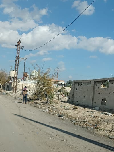 The image depicts a daytime scene on a dirt or heavily worn asphalt road in Al-Qadam Municipality, Syria, under a blue sky with scattered white clouds. The sun casts a long, dark shadow diagonally across the road from left to right.

In the mid-ground, a solitary individual, appearing male, walks away from the camera on the right side of the road's shoulder. He is dressed in a light brown jacket, blue jeans, and a red and white checkered keffiyeh draped around his head and neck. A light-colored bag or sack is slung over his left shoulder.

To the left of the person stands a tall, multi-component utility pole constructed of rusted metal, featuring cross arms and insulators, supporting overhead power lines. A heavy black cable extends from the pole towards the upper right of the frame. Beyond the utility pole and the individual, several buildings are visible. One prominent structure features a light-colored dome, indicative of a mosque or similar religious building. Other structures appear to be in various states of repair or disrepair.

To the right of the walking individual and extending towards the right edge of the frame, a concrete or masonry wall stands severely damaged. This wall exhibits numerous bullet holes and larger impact craters, with some sections completely broken through. Its top edge is irregular, and an empty window aperture is visible. The ground immediately adjacent to the road and in front of the damaged wall is unkempt, littered with debris, loose earth, and sparse, low-lying vegetation and bushes. The overall environment suggests a semi-urban area affected by conflict or neglect.