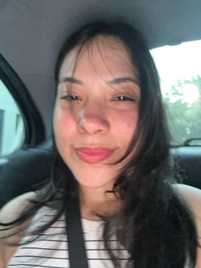 A female individual is centrally positioned within the interior of a vehicle, facing the camera. She has long, dark hair, with some strands partially obscuring her face, and displays a slight smile with pink-colored lips. Her eyes are dark and appear slightly narrowed. She is wearing a light-colored garment with horizontal dark stripes, and a dark strap is visible across her upper chest. The vehicle's dark-colored interior forms the immediate background. Through the left window, a light-colored structure is visible, while the right window reveals blurred green foliage and bright outdoor light. The scene is situated in Villa Yapeyú, Argentina.