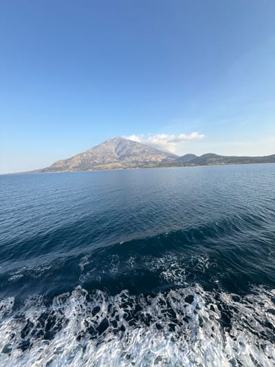 The image depicts a seascape with the coastline of Samothraki island visible from the sea, specifically from the vicinity of Kamariotissa, Greece. The foreground features turbulent dark blue water with a pronounced white, foamy wake extending from the bottom edge and receding into the midground, indicating the movement of a vessel. The midground consists of calmer, deep blue water. The background is dominated by a large, rugged mountain, Mount Fengari, which rises directly from the coastline. Its slopes are characterized by rocky outcrops and patches of sparse green vegetation. Small clusters of buildings and cultivated green areas are discernible along the mountain's base, particularly on the right side of the frame, consistent with coastal settlements. The sky above is clear and light blue, with a few wispy white clouds clustered near the mountain peak on the right. No individuals or additional man-made objects are present.