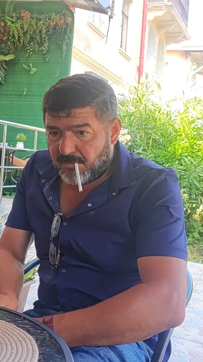 A man with dark hair, a beard, and a mustache is sitting at a table outdoors. He is wearing a navy blue collared shirt and blue jeans. A cigarette is held in his mouth. His eyes are looking down and to the left. A pair of eyeglasses is hanging from the collar of his shirt. Behind him, there is lush green foliage and a building with arched windows. The lighting suggests it is daytime, possibly late morning or early afternoon, with bright sunlight. The setting appears to be a cafe or restaurant patio in Dagomys, Russia, given the visible greenery and architectural style. The man's expression is neutral. No text is visible in the image.