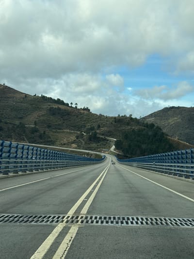 The image displays a four-lane highway, likely a viaduct or bridge, observed from within a moving vehicle in Carrazeda de Ansiães, Portugal. In the foreground, a metal expansion joint or drainage grate spans the width of the asphalt road, which is marked with double solid yellow lines separating directions and dashed white lines for lanes. Blue, modular crash barriers featuring a geometric pattern line both sides of the highway, curving gently to the right as the road descends into a valley and subsequently ascends into the distance. Two white passenger vehicles are visible in the far midground, traveling away from the viewer in the rightmost lane. The surrounding terrain comprises steep, terraced hillsides on the left and rugged, vegetated hills on the right, both featuring scattered trees and visible winding roads. The sky is predominantly overcast with large grey clouds, though a distinct patch of blue sky with white clouds is visible in the upper right quadrant.