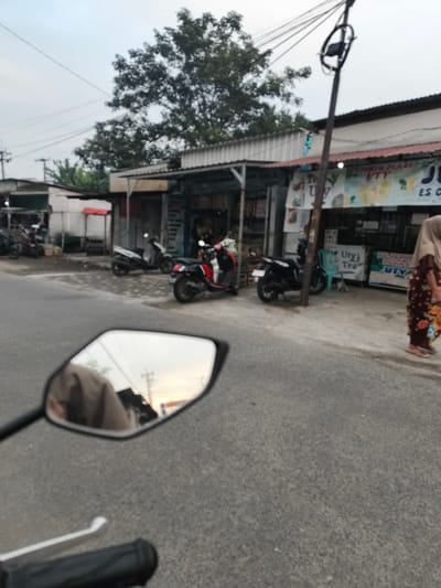 A street-level, slightly blurred photograph depicts a row of small shops lining a paved road in Sidorejo, Indonesia. The shops appear to be typical Indonesian roadside businesses, with awnings and open fronts displaying goods. Several motorcycles are parked or displayed in front of the shops. One red and black scooter is prominent on the right, and a white and black scooter is parked further back. A woman wearing a hijab and a patterned dress walks away from the shops on the sidewalk. In the foreground, a motorcycle's rearview mirror is visible, reflecting the sky and part of the scene. The overall atmosphere suggests a commercial area during daylight, possibly late afternoon or early morning given the lighting. A prominent utility pole with many wires stands near the shops.
