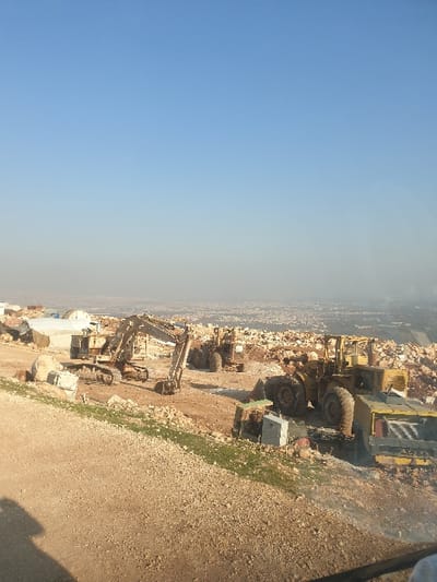 The image displays an extensive outdoor industrial site, likely a quarry or a large-scale construction area, located in Killi, Syria. The scene is dominated by rugged terrain covered with vast quantities of earth, gravel, and large, irregular rocks and boulders.
Several pieces of heavy machinery are visible across the site. Prominently positioned on the right side of the frame is a large yellow front-end loader with substantial tires, suggesting its use in moving heavy materials. Further into the mid-ground, another large yellow piece of equipment, possibly another loader or a specialized truck, is partially visible. Towards the left in the mid-ground, a large yellow excavator with tracks is situated amidst piles of rocky debris, indicating excavation work. Various other smaller structures and equipment, including some possibly covered in white tarps, are scattered throughout the site.
The sky above is clear and bright blue, transitioning to a lighter, hazy shade closer to the horizon. In the far distance, a faint, low-lying landscape with hints of buildings or a settlement can be seen, suggesting a populated area beyond the immediate industrial zone. Sparse patches of green vegetation are visible on the ground, especially along the edges of the disturbed earth.
No people are present in the image. The environment strongly suggests ongoing or recently completed activities related to quarrying, earthmoving, or large-scale construction. The time of day appears to be bright daylight, under clear weather conditions. There is no visible text in the image.