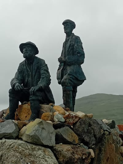Two bronze statues of men stand on a pile of rocks under an overcast sky. The statue on the left depicts a man with a beard wearing a hat and a jacket, seated with his hands resting on his knees. The statue on the right shows a man wearing a cap and a jacket, standing with his hands clasped in front of him. In the background, rolling green hills are visible. The scene appears to be outdoors in a rugged, natural setting.